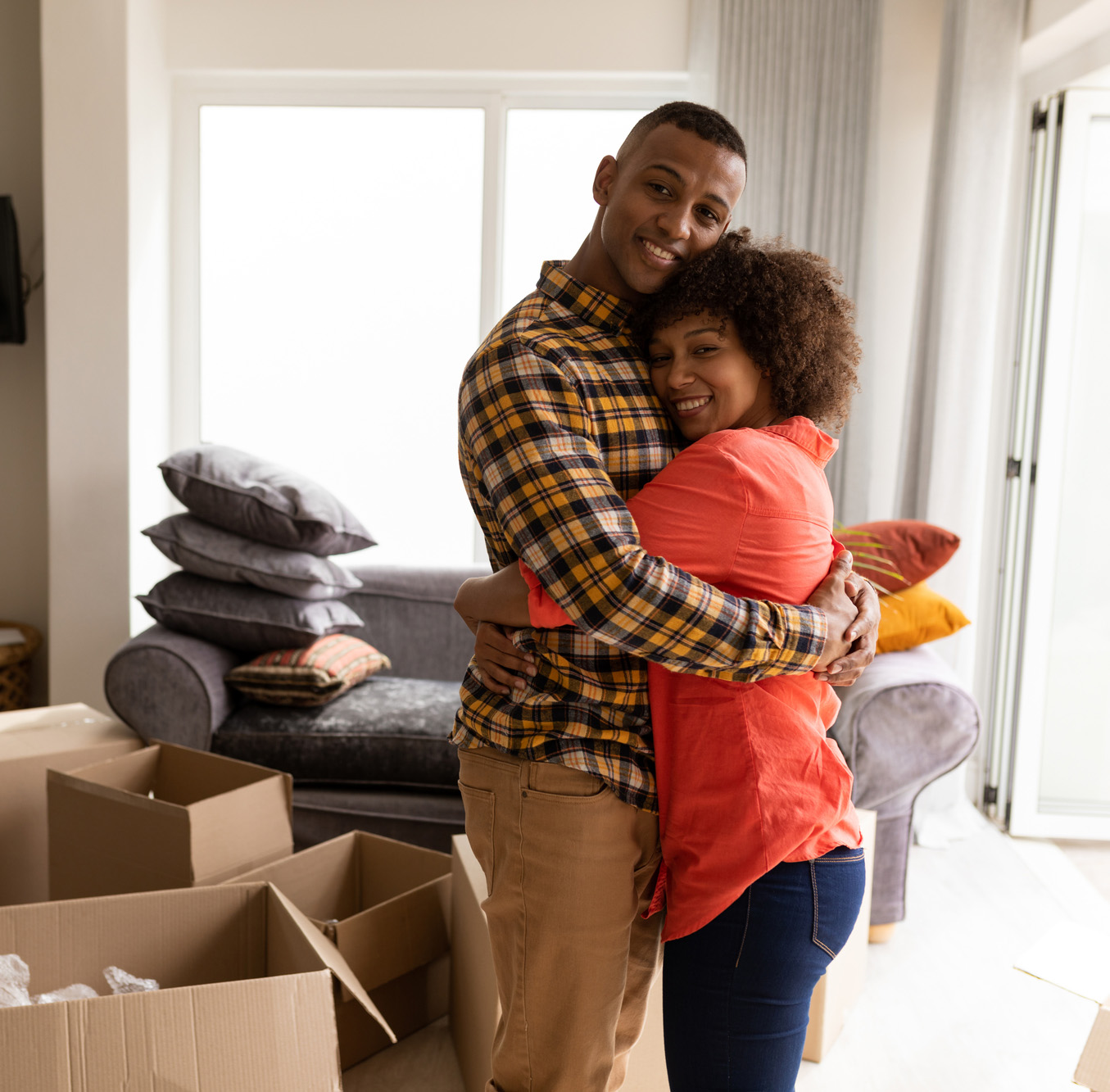 Portrait of happy young mixed-race couple embracing each other in living room at home. They are surrounded by cardboard boxes.
